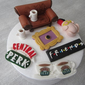 May include: A white cake decorated with fondant figures and text. The cake features a brown couch, a purple picture frame, a turkey wearing a fez, two coffee mugs, and two signs that say "Central Perk" and "F.R.I.E.N.D.S."