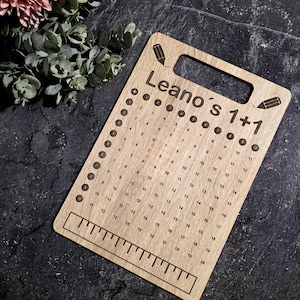 Könnte beinhalten: Eine hölzerne Multiplikationstafel mit dem Text "Leano's 1+1" und einem Lineal am unteren Rand. Die Tafel hat einen Griff und Bleistiftillustrationen. Zahlen von 1 bis 20 sind in Zeilen und Spalten angeordnet.