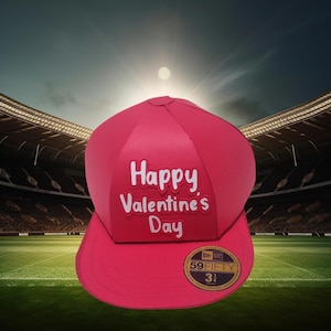 Puede incluir: Gorra de béisbol rosa brillante con el texto "Happy Valentine's Day" en blanco. La gorra tiene una visera curva y una pegatina de talla 7 3/4. El fondo muestra un estadio con un campo verde y un cielo brillante.