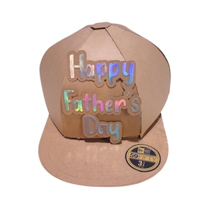 Puede incluir: Un sombrero de cartón marrón con forma de gorra de béisbol con las palabras "Happy Father's Day" en letras iridiscentes en la parte delantera. El sombrero tiene una pegatina New Era 59FIFTY negra y dorada en el lateral.