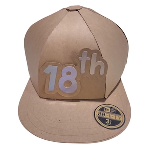 Puede incluir: Una gorra de béisbol de cartón marrón con una pegatina 18th plateada y blanca en la parte delantera. La gorra tiene una pegatina New Era 59FIFTY negra y dorada en el lateral.