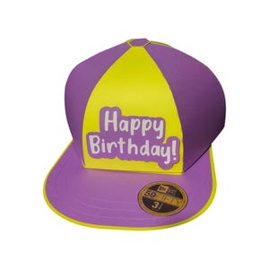 Puede incluir: Gorra de béisbol morada y amarilla con el texto "Happy Birthday!". La gorra tiene una visera curva y una pegatina 59FIFTY. Un accesorio festivo para una celebración de cumpleaños.
