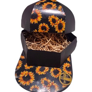 Puede incluir: Caja de regalo negra y amarilla con estampado de girasoles con forma de gorra de béisbol. La caja está abierta y tiene un relleno de papel marrón en el interior. La caja tiene una pegatina dorada que dice "59FIFTY 3 1/8"