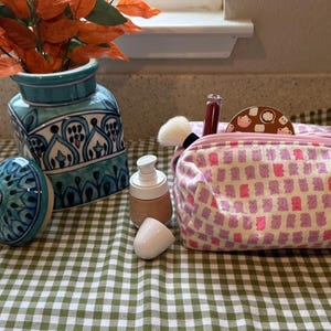 May include: A pink and white patterned cosmetic bag with a zipper closure. The bag is filled with makeup brushes, a bottle of lotion, and a small compact. The bag is sitting on a green and white checkered tablecloth.