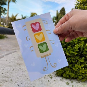 Può includere: Una cartolina tenuta in mano, con un design stravagante di semaforo. Il semaforo ha luci a forma di cuore in rosso, giallo e verde. Lo sfondo è azzurro con piccole forme di cuore. La cartolina ha una base decorativa e vorticosa.