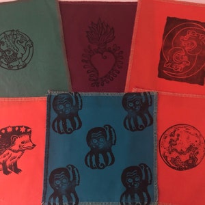 Op de afbeelding: Een verzameling kleurrijke stoffen vierkanten met diverse zwarte linosnede ontwerpen. De ontwerpen omvatten een griffioen, een hart met vlammen, een maan met figuren, een egel, octopusachtige wezens en een volle maan.