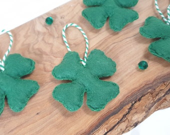 May include: Five green felt four-leaf clover ornaments with white and green twine hanging from a wooden board.