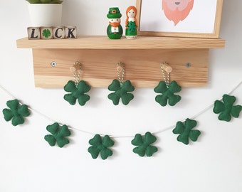 May include: A green felt shamrock garland with white string hanging from a wooden shelf. The garland has nine shamrocks and is decorated with two small figurines of a leprechaun and a woman.