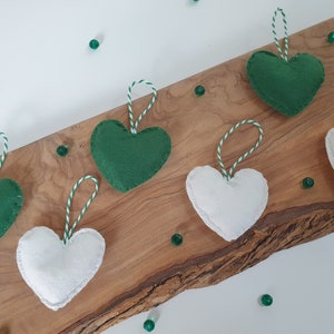 May include: Six felt heart ornaments, three green and three white, with white and green twine hanging from each. The ornaments are displayed on a wooden surface.