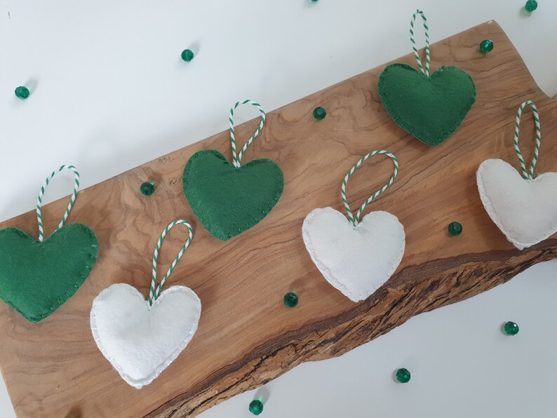 May include: Six felt heart ornaments, three green and three white, with white and green twine hanging from each. The ornaments are displayed on a wooden surface.