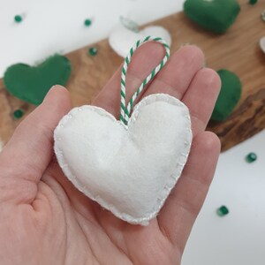 May include: A white felt heart ornament with a green and white striped string for hanging. The heart is stitched around the edges.