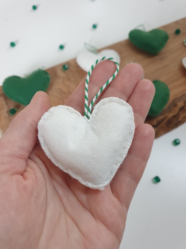 May include: A white felt heart ornament with a green and white striped string for hanging. The heart is stitched around the edges.