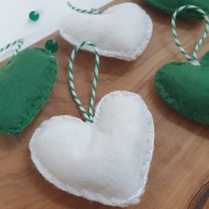 May include: Five felt heart ornaments, two green and three white, with white and green twine attached. The hearts are on a wooden surface.