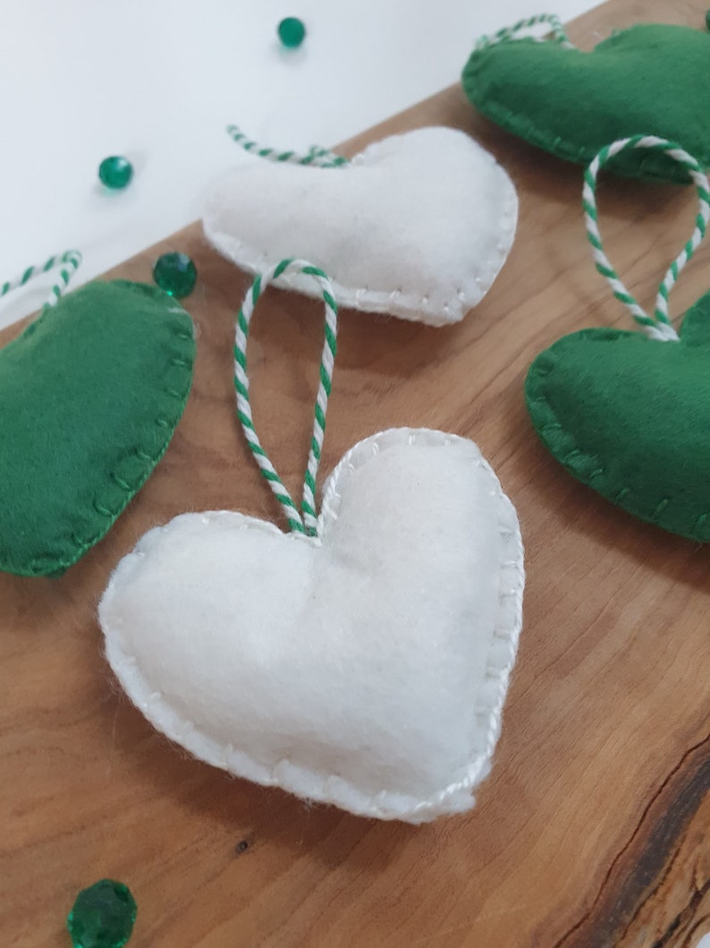 May include: Five felt heart ornaments, two green and three white, with white and green twine attached. The hearts are on a wooden surface.