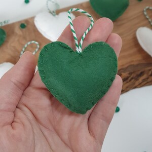 May include: A green felt heart ornament with white stitching around the edges. The ornament is hanging from a white and green twine string.