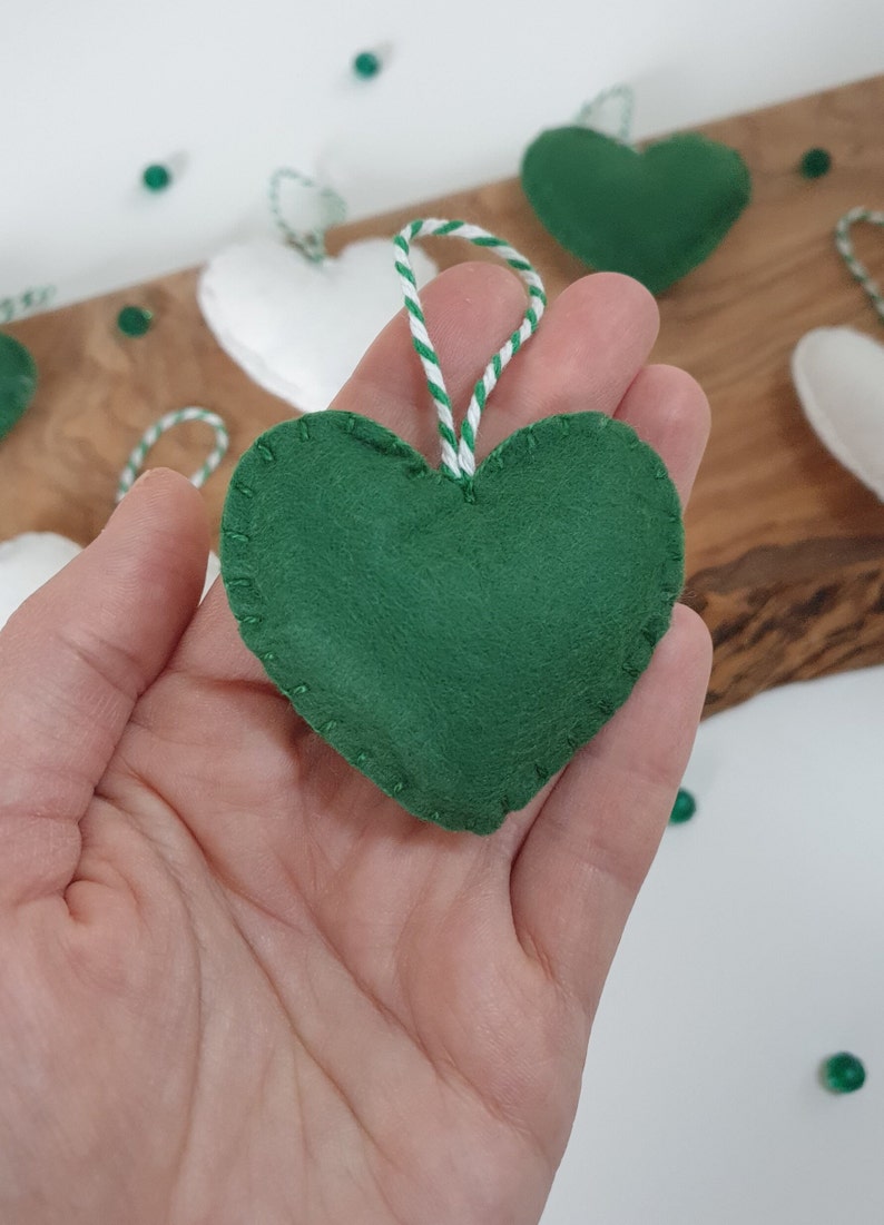 May include: A green felt heart ornament with white stitching around the edges. The ornament is hanging from a white and green twine string.
