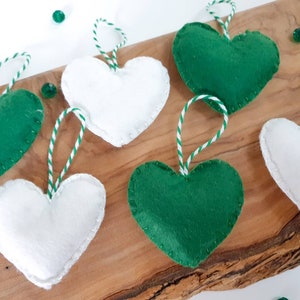 May include: Six felt heart ornaments, three green and three white, with white and green twine hanging loops. The ornaments are arranged on a wooden surface.