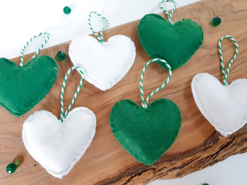 May include: Six felt heart ornaments, three green and three white, with white and green twine hanging loops. The ornaments are arranged on a wooden surface.