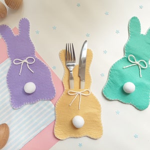 May include: Three felt bunny-shaped utensil holders. The bunny on the left is purple, the bunny in the middle is yellow, and the bunny on the right is green. Each bunny has a white pom-pom tail and a white ribbon tied around its neck.