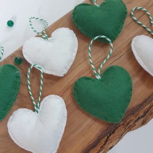 May include: Six felt heart ornaments, three white and three green, with white and green twine attached. The ornaments are arranged on a wooden surface.