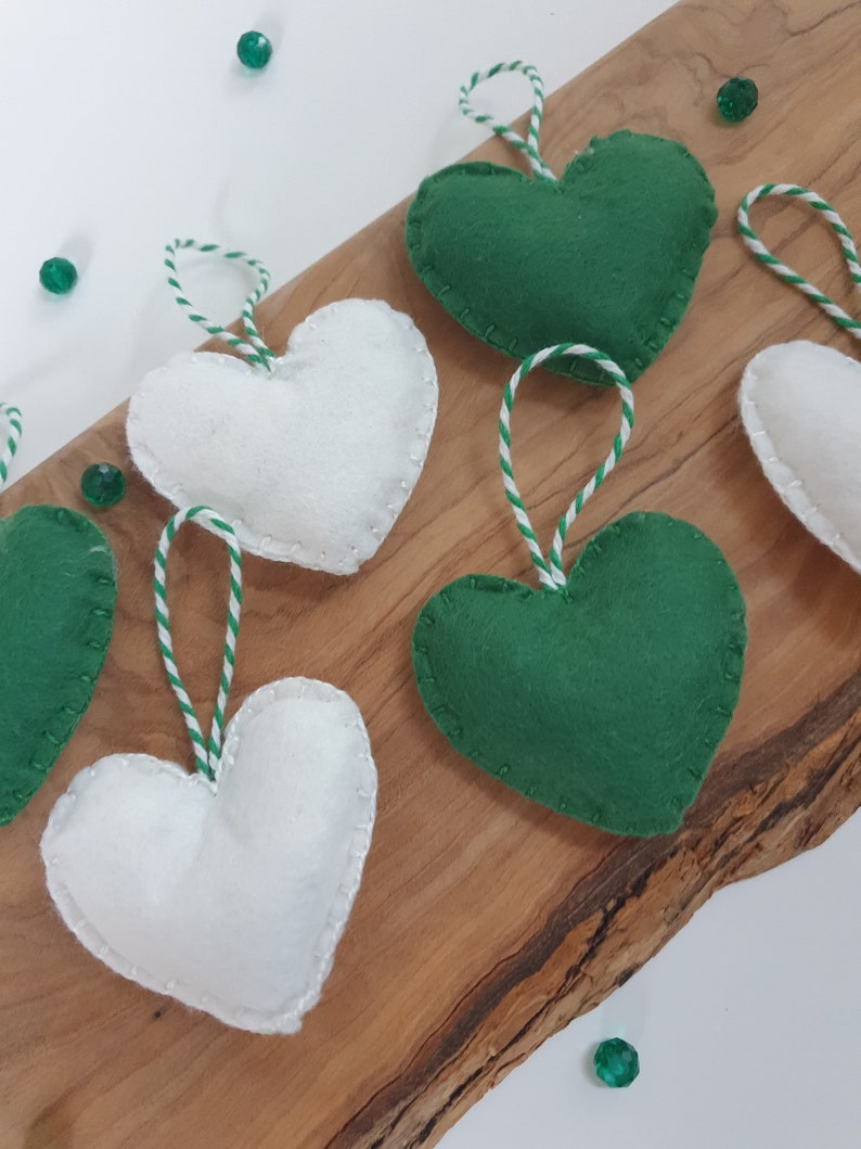 May include: Six felt heart ornaments, three white and three green, with white and green twine attached. The ornaments are arranged on a wooden surface.