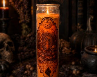 Op de afbeelding: Een hoge, cilindrische glazen kaars gevuld met oranje was, voorzien van een etiket met een pompoen, een schedel en het silhouet van een zwarte kat. De tekst op het etiket luidt SAMHAIN en Black Cat Curiosity Co. De kaars brandt met een zichtbare vlam.