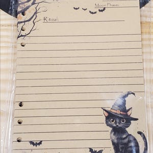 Op de afbeelding: Een bruine papieren journalpagina met een zwarte kat die een heksenhoed draagt. De pagina heeft lijnen om op te schrijven en bevat de tekst "Ritueel:", "Datum:" en "Maanfase:". Er zijn ook zwarte vleermuizen getekend op de pagina.