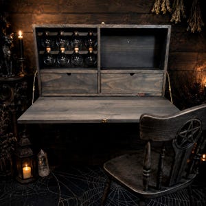 May include: A wall-mounted dark wood drop-leaf desk featuring a fold-down work surface, two drawers, and an upper storage section with eight glass vials with cork stoppers. A matching wooden chair with a carved pentagram backrest is positioned below.