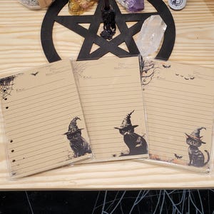 Op de afbeelding: Drie bruine papieren notitieboekjes met zwarte kattenillustraties die toverhoeden dragen. Elk notitieboekje heeft de titel "Ritual" en ruimte om de datum, maand en jaar te schrijven.