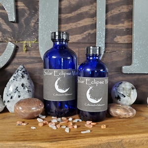 May include: Two blue glass bottles with black labels that say "Solar Eclipse Water Collected in Missouri" and a crescent moon graphic. The bottles are sitting on a wooden surface with small, colorful stones.