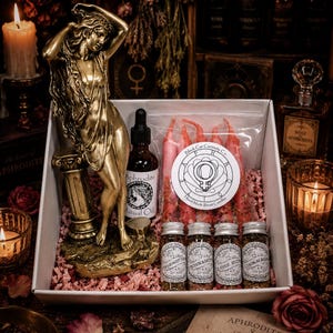 May include: A gift box containing a gold-toned statue of a figure leaning on a pillar, a bottle of Aphrodite ritual oil, a plastic bag of five red ritual candles with a circular label, and four small glass jars filled with dried herbs.
