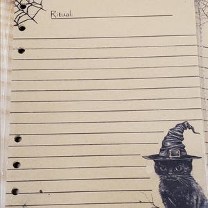 Op de afbeelding: Een lege pagina uit een heksendagboek met een zwarte kat die een heksenhoed draagt in de rechterbenedenhoek. De pagina heeft lijnen om op te schrijven en een spinnenwebontwerp in de linkerbovenhoek. De tekst "Ritueel:" staat bovenaan de pagina. De tekst "Datum:" en "Maanfase:" staat rechtsboven.