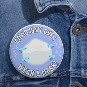 May include: A blue button with the text "COVID ISN'T OVER WEAR A MASK!" and a white face mask with blue straps. The button has a white border and a white background with blue stars.