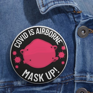May include: A black and white button with the text "COVID IS AIRBORNE MASK UP!" in white. A pink face mask with black hearts is in the center of the button. There are red stylized coronavirus images around the mask.