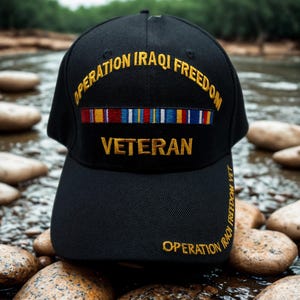 May include: Black baseball cap with gold embroidered text reading "OPERATION IRAQI FREEDOM VETERAN." The cap features a row of colorful ribbons and the words "OPERATION IRAQI FREEDOM VET" embroidered on the side. The cap is sitting on a bed of rocks.