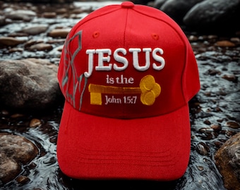 May include: A red baseball cap with the text "JESUS is the John 15:7" embroidered in white and gold. The cap features a curved brim and a structured crown. The background shows a stream with rocks.