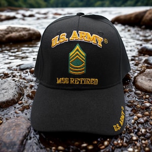 Puede incluir: Gorra de béisbol negra con bordado dorado "U.S. ARMY". La gorra presenta una insignia dorada y verde y el texto "MSG RETIRED". La gorra está sobre un fondo de rocas y agua.