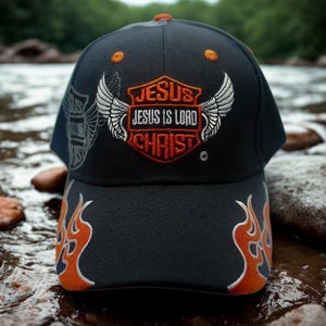 Może przedstawiać: Czarna czapka z daszkiem z pomarańczowymi i białymi haftowanymi płomieniami na rondzie. Z przodu znajduje się wzór tarczy ze słowami "JESUS IS LORD CHRIST" i skrzydłami w kolorze pomarańczowym i białym.