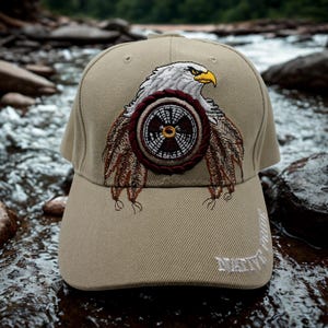 May include: A tan baseball cap featuring an embroidered design of a white eagle head with a yellow beak, positioned above a circular emblem with brown and white patterns. The text NATIVE PRIDE is embroidered in white on the lower right brim.