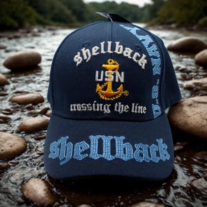 Peut inclure: Casquette de baseball bleu marine avec texte brodé et un emblème d'ancre dorée. La casquette présente les mots "Shellback", "USN" et "crossing the line" en blanc et bleu clair. Un accessoire élégant pour les activités de plein air.