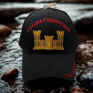 May include: Black baseball cap with "COMBAT ENGINEER" embroidered in red and a gold castle emblem. The cap is made of a durable fabric and features a curved brim. The hat is displayed in a natural outdoor setting.