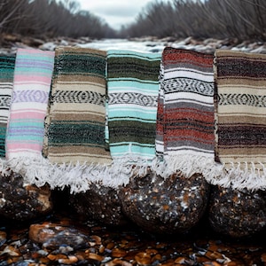 May include: A collection of six folded woven blankets featuring various color combinations including teal, pink, green, and brown, each with a distinct striped pattern and white fringed edges along the bottom, displayed in a row.