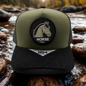 May include: A black and olive green trucker hat featuring a horse head patch. The patch is circular with a horse head silhouette and the word "HORSE". The hat has a mesh back and a curved brim. The text "PREMIUM QUALITY HEADWEAR 804 KAMEL" is printed on the brim.