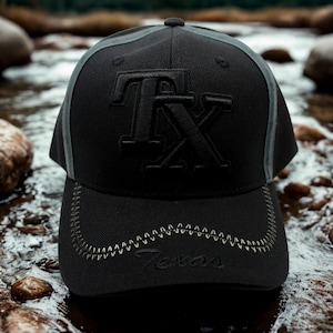May include: Black baseball cap featuring embroidered "TX" lettering. The brim has "Texas" stitched on it. The cap has a grey accent stripe and a curved brim with decorative stitching.