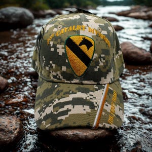 May include: Camouflage baseball cap with the text "1st Cavalry Division" and a patch featuring a black horse on a yellow and black shield. The cap also has the text "THE FIRST TEAM!" in gold.