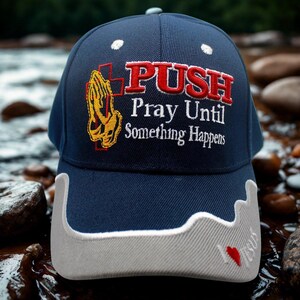 May include: A navy blue baseball cap with white detailing and embroidered text. The cap displays the words "PUSH Pray Until Something Happens" in red and white, alongside a gold and orange praying hands graphic. A small red heart is embroidered on the brim.
