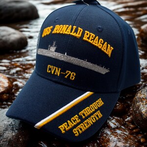 Op de afbeelding: Marineblauwe baseballcap met goudkleurig geborduurde tekst en een scheepsillustratie. De cap heeft "USS RONALD REAGAN" en "CVN-76" op de voorkant, met "PEACE THROUGH STRENGTH" op de klep. Een stijlvol accessoire.
