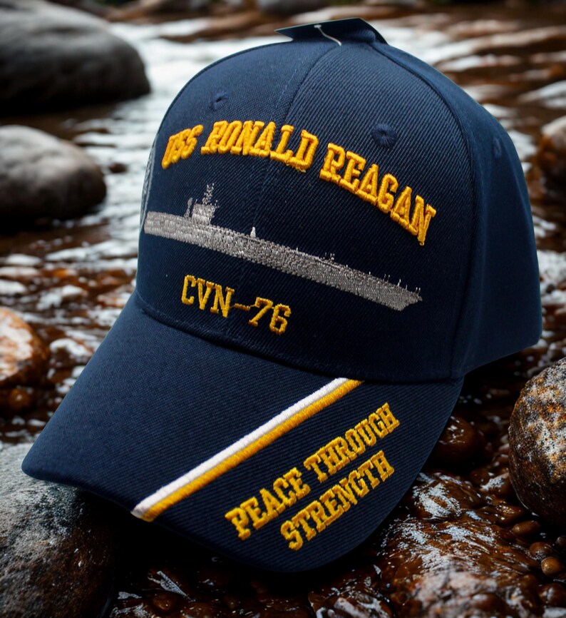 Op de afbeelding: Marineblauwe baseballcap met goudkleurig geborduurde tekst en een scheepsillustratie. De cap heeft "USS RONALD REAGAN" en "CVN-76" op de voorkant, met "PEACE THROUGH STRENGTH" op de klep. Een stijlvol accessoire.