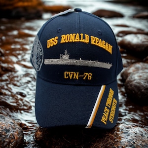 Op de afbeelding: Marineblauwe baseballcap met gouden borduursel met de tekst "USS Ronald Reagan" en "CVN-76". De cap heeft ook een scheepsillustratie en de woorden "Peace Through Strength" in goud.