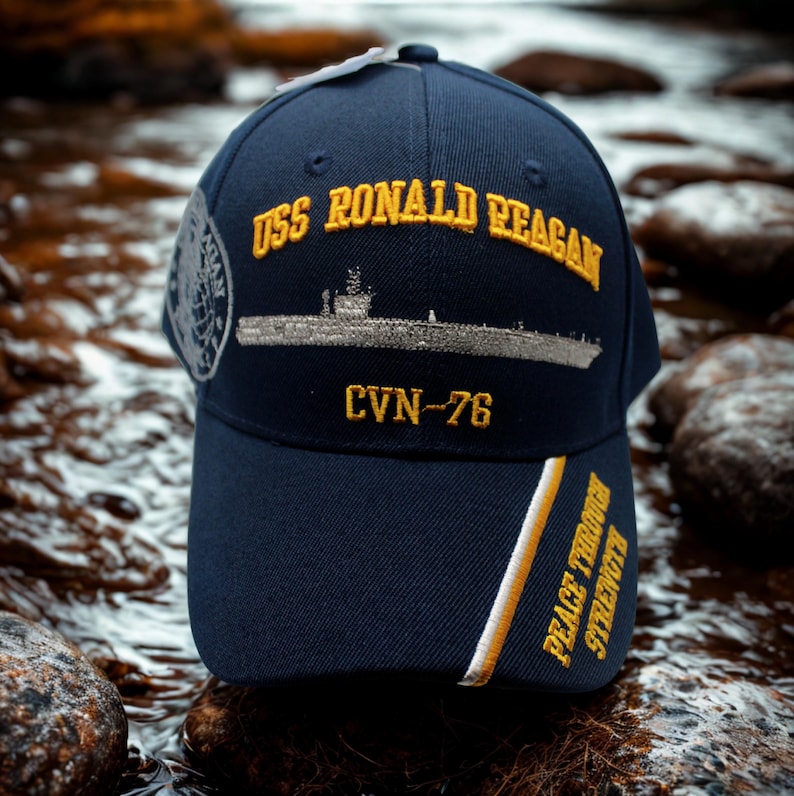 Op de afbeelding: Marineblauwe baseballcap met gouden borduursel met de tekst "USS Ronald Reagan" en "CVN-76". De cap heeft ook een scheepsillustratie en de woorden "Peace Through Strength" in goud.